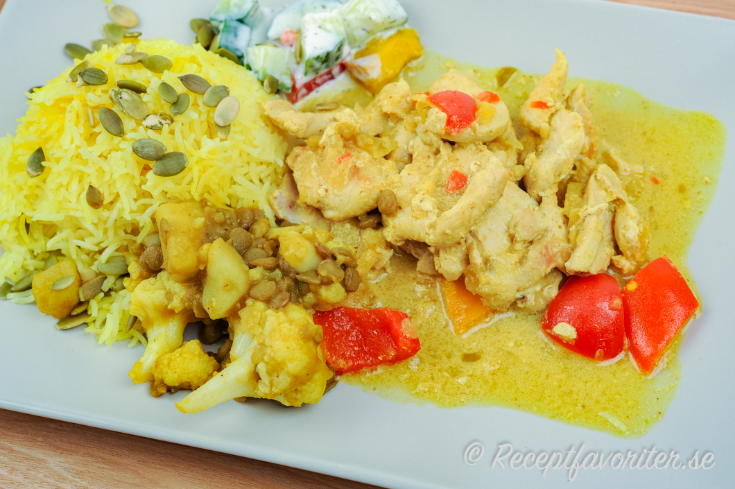 Gryta med kycklingfilé och curry serverad med tillbehör som raita, ris och linser. 