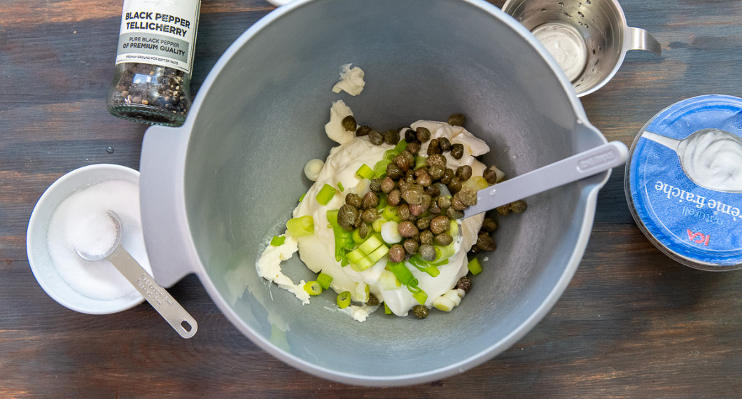 Mät upp crème fraiche, majonnäs, salladslök och kapris i en skål. 