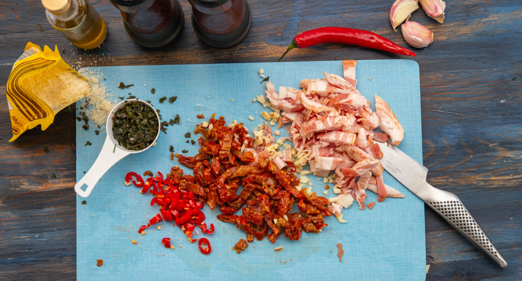 Strimlad bacon, hackade soltorkade tomater, vitlök och chili på skärbräda. 