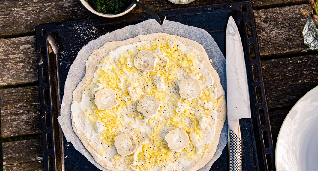 Pizzabotten förbereds med ett lager crème fraiche, riven mozzarella, smulad färsk mozzarella samt chèvre. 
