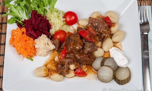 Gulasch serverad med skivad saltgurka, gräddfil och kokt gnocchi samt en sallad av grovrivna råa rödbetor, finrivna råa morötter, finriven rå rotselleri, grovriven zucchini, cocktailtomater samt grönsallad.
