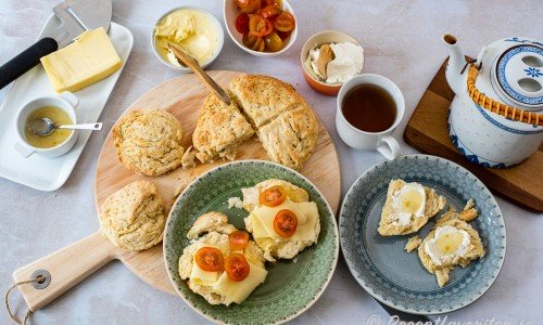 Scones serverad med te och tillbehör som marmelad, ost, smör, tomat och färskost. 