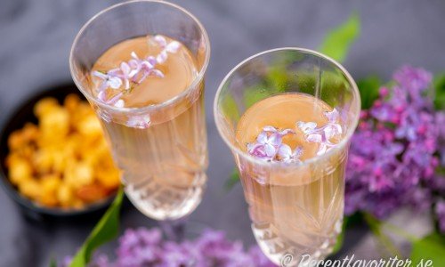 Två bubblande glas Syren Prosecco garnerade med syrenblommor. 