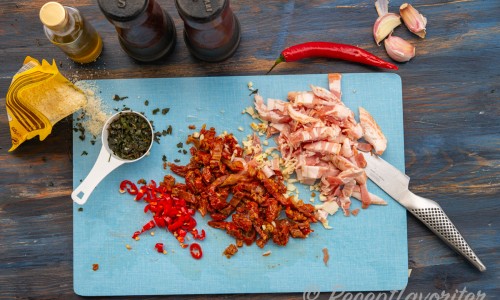 Strimlad bacon, hackade soltorkade tomater, vitlök och chili på skärbräda. 
